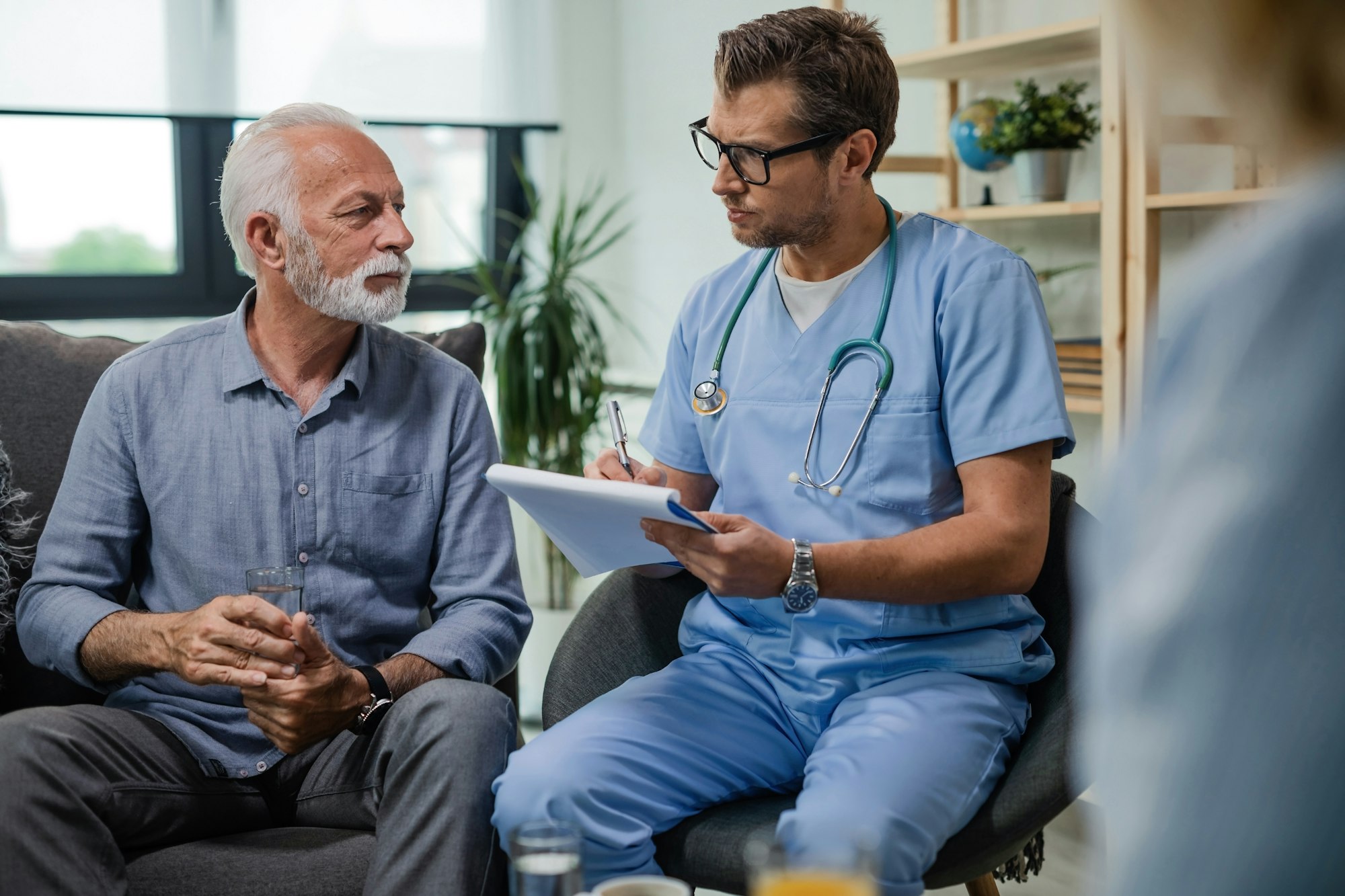 General practitioner taking notes and communicating with senior patient while being in home visit.